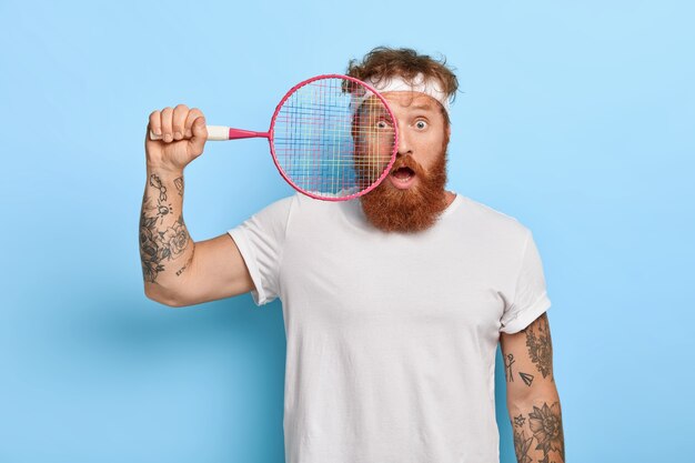 Horizontal shot of scared red haired tennis player holds racket while posing against the blue wall