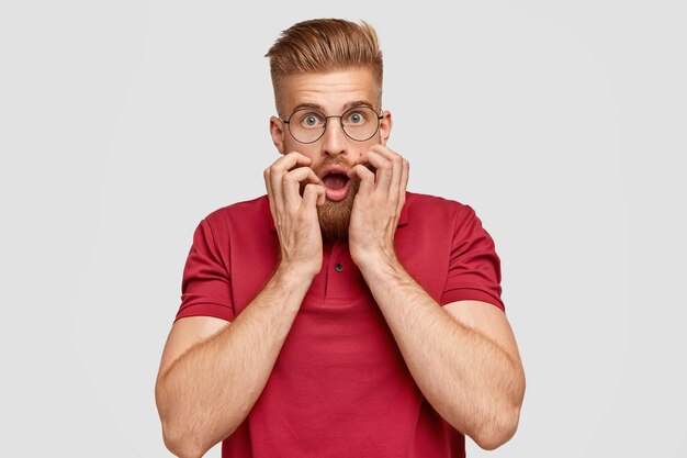 Horizontal shot of scared stunned man with foxy beard keeps both hands near mouth