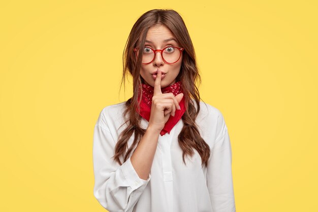 Horizontal shot of secret girl makes shush gesture, keeps fore finger over lips, asks not spread rumors, poses against yellow wall. People, secrecy, conspiracy concept. People and body language