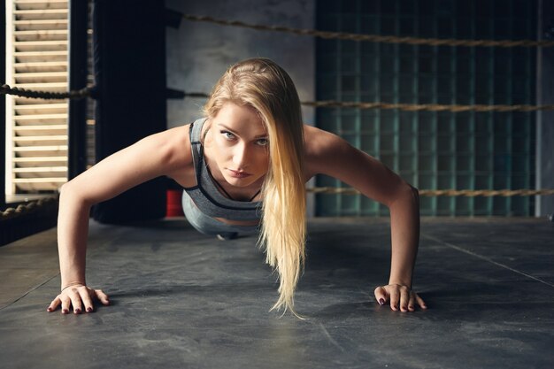 Horizontal shot of self determined gorgeous young sportswoman with loose dyed hair doing push ups planting hands widely on floor inside boxing ring