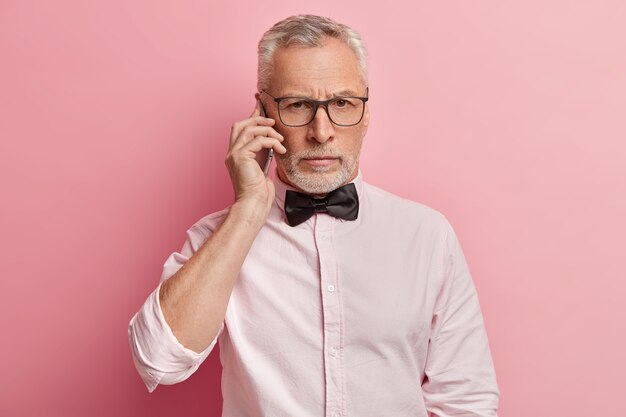 Horizontal shot of senior man talks on phone with serious expression, holds modern mobile phone near ear, has conversation