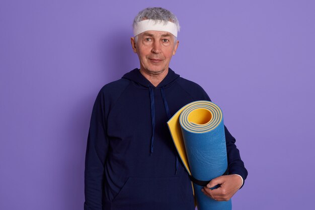 Horizontal shot of senior white haired male with head band and holding blue yoga mat in hands