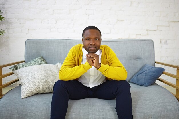 Горизонтальный снимок серьезного взрослого афро-американского мужчины в стильной одежде, сидящего на сером диване со сложенными руками, с задумчивым выражением лица, думающего о чем-то. Люди и образ жизни