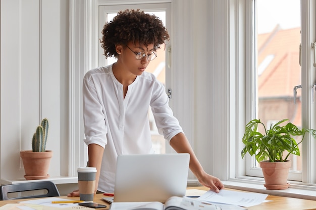 Horizontal shot of serious black young woman stands at desktop