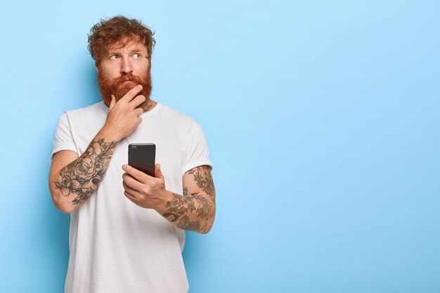 Horizontal shot of serious contemplative adult man touches thick red beard, holds mobile phone, browses newsfeed online, thinks over recent news, has tattooed arms, wears casual white t shirt