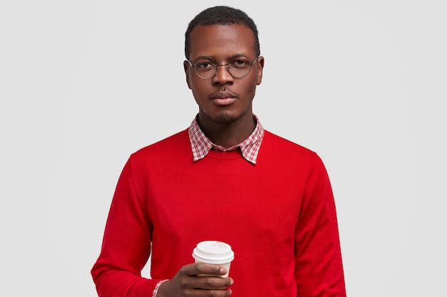 Horizontal shot of serious dark skinned man holds disposable cup of coffee, dressed in red jumper, looks confidently 