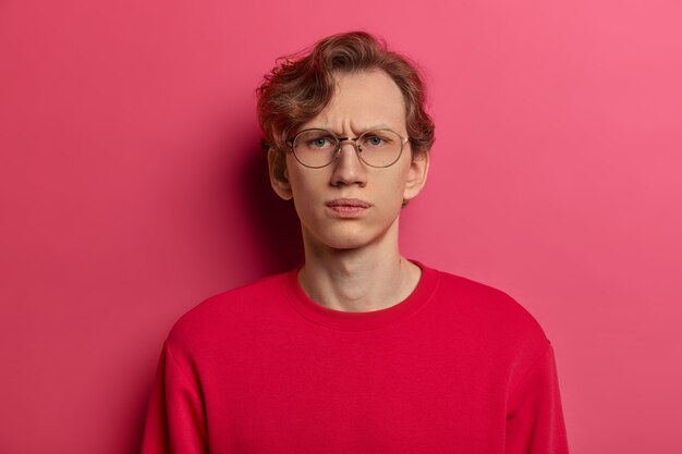 Horizontal shot of serious dissatisfied male model smirks face and looks straightly , doubts he can trust you, wears spectacles and red sweater, isolated on pink wall, feels intense