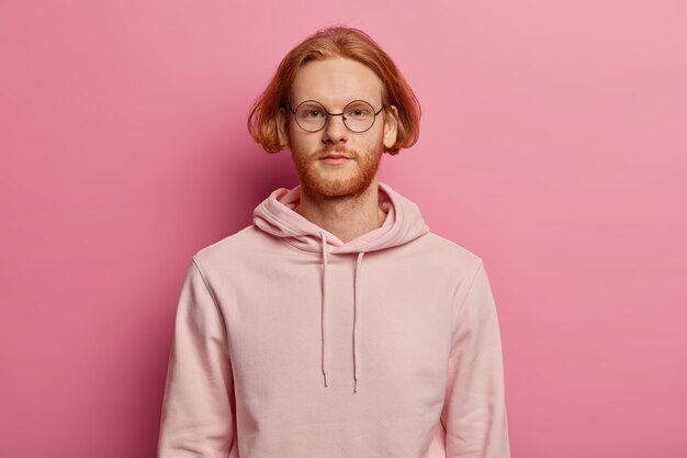 Horizontal shot of serious looking bearded European man looks directly , has calm expression, wears casual hoodie, poses against rosy wall, talks to someone, has bob hairstyle