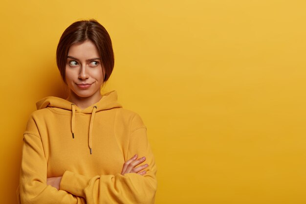 Horizontal shot of serious looking European woman crosses hands over chest, thinks about something with doubt, wears casual hoodie, poses against yellow  wall, doesnt want to talk to you