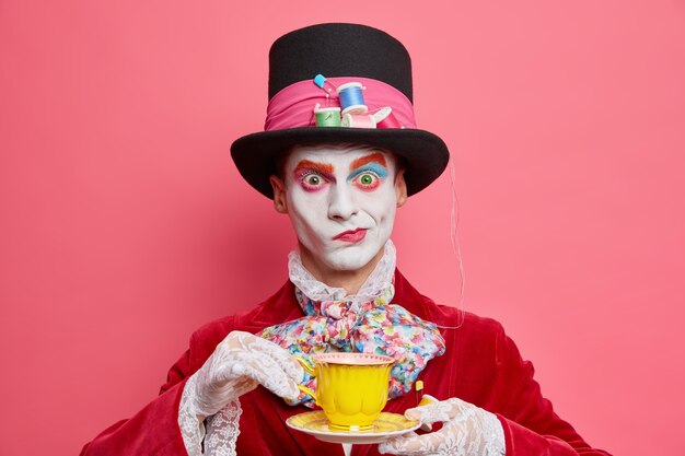 Horizontal shot of serious male hatter poses with cup of tea wears hat has manners of aristocratic gentleman dresses for masquerade carnival poses indoor