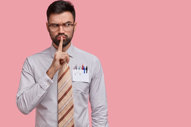 Horizontal shot of serious male office worker dressed in formal shirt and tie, demonstrates shush sign