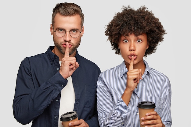 Horizontal shot of serious mixed race female and male keep index fingers over mouth, make shush gesture