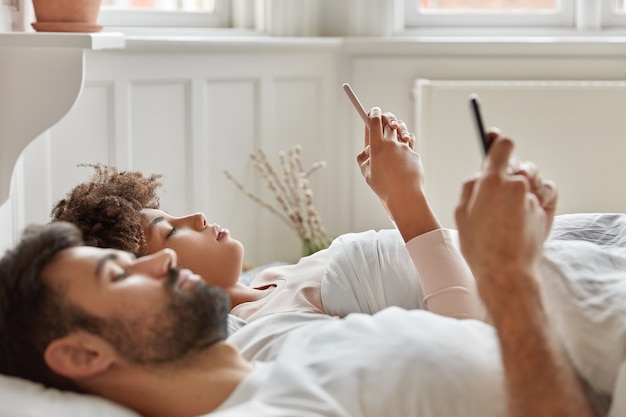 Horizontal shot of serious multiethnic young woman and man focused in modern gadgets, monitor web multimedia content