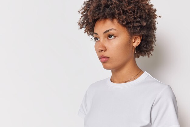 Horizontal shot of serious thoughtful young woman focused away with pensive expression considers something wears casual t shirt poses against white background blank space for your promotion.