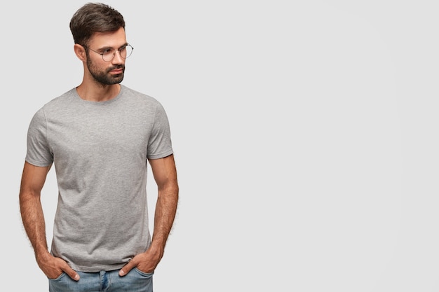 Горизонтальный снимок серьезного небритого мужчины в повседневной серой футболке, держит руки в карманах, смотрит в сторону, о чем-то думает