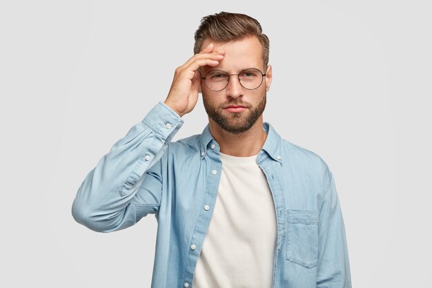 Horizontal shot of serious unshaven male has pensive expression, keeps hand on forehead, tries to gather with thoughts, dressed in blue shirt, being intelligent, isolated over white wall