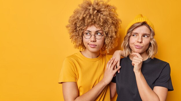 Horizontal shot of serious young women focused away and ponder on something have thoughtful expressions dressed in casual t shirts pose against yellow background copy space for your promotion