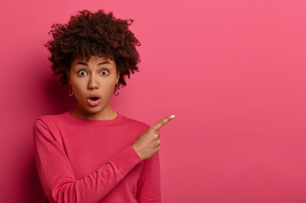 Horizontal shot of shocked dark skinned curly haired woman keeps mouth opened, gasps from wonder, demonstrates something with low price, wears pink jumper, gestures indoor. Advertisement concept