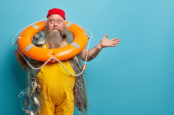 Free Photo horizontal shot of shocked embarrassed bearded male seafarer poses with inflated lifering fishing net smokes pipe raises hand over blue wall demonstrates copy space for your advertising content