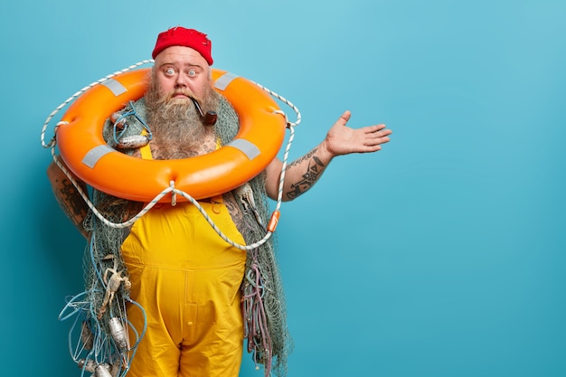 Горизонтальный снимок потрясенного, смущенного бородатого моряка-мужчины позирует с надутой жизнерадостной рыболовной сетью, курит трубку, поднимает руку над синей стеной, демонстрирует место для копирования вашего рекламного контента