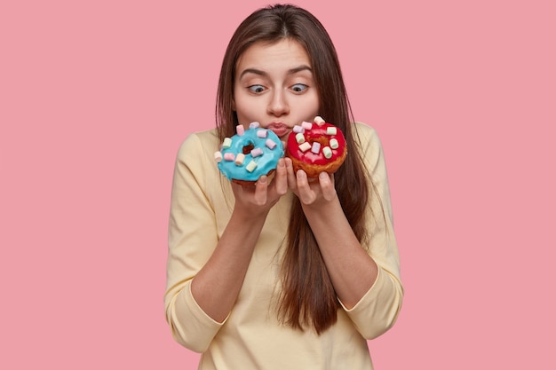 Horizontal shot of shocked pretty European woman holds blue and red doughnuts, smells aromatic confectionery