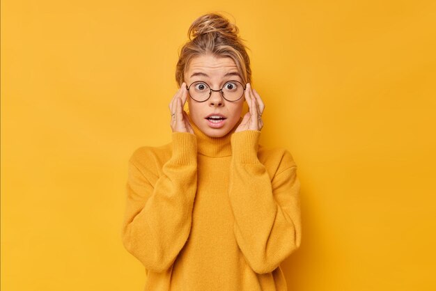 Horizontal shot of shocked puzzled woman holds breath cannot believe her eyes keeps hands on rim of spectacles feels stupefied wears sweater round spectacles isolated over yellow background.