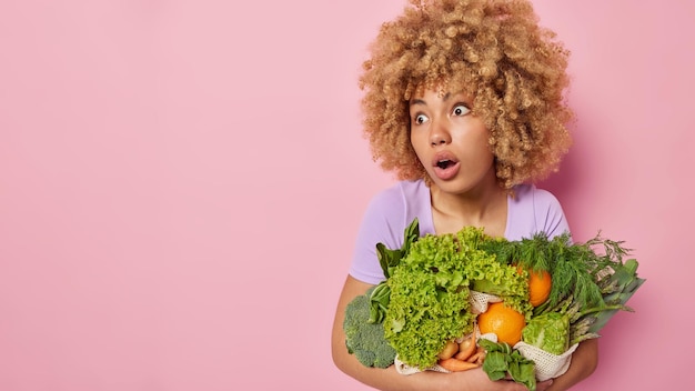 Foto gratuita l'inquadratura orizzontale di una donna scioccata con i capelli ricci e folti mantiene la bocca aperta non può credere che i propri occhi portino verdure fresche primaverili coltivate in serra isolate su sfondo rosa spazio vuoto