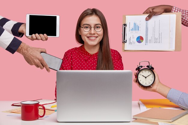 Horizontal shot of smiling beautiful woman in red shirt, sits in front of opened laptop computer