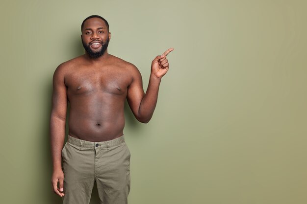 Horizontal shot of smiling black bearded man poses with bare torso indicates at empty space