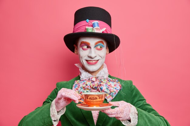 Horizontal shot of smiling cheerful mysterious hatter spends free time on tea party wears hat and costume with bowtie has holiday mood poses against pink wall