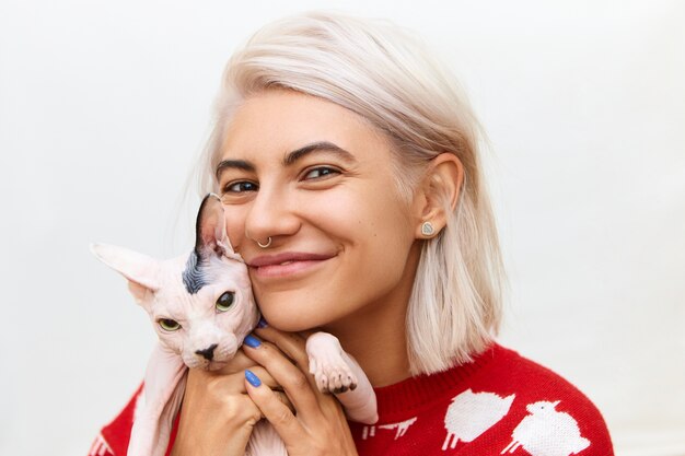 Horizontal shot of smiling pretty girl with nose ring and bob hairstyle spending time with her pet, embracing gray Sphynx cat tight, showing love, care, having joyful happy facial expression