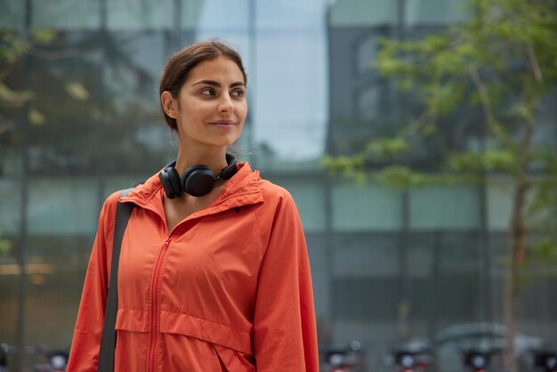 Horizontal shot of sporty brunette woman dressed in windbreaker carries rolled karemat listens music via headphones poses outdoors against blurred background Aerobics and active lifestyle concept
