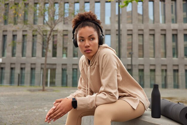 Horizontal shot of sporty thoughtful curly haired woman focused away dressed in sportswear listens music via headphones sits outdoors near bottle of water and fitness mat against city building