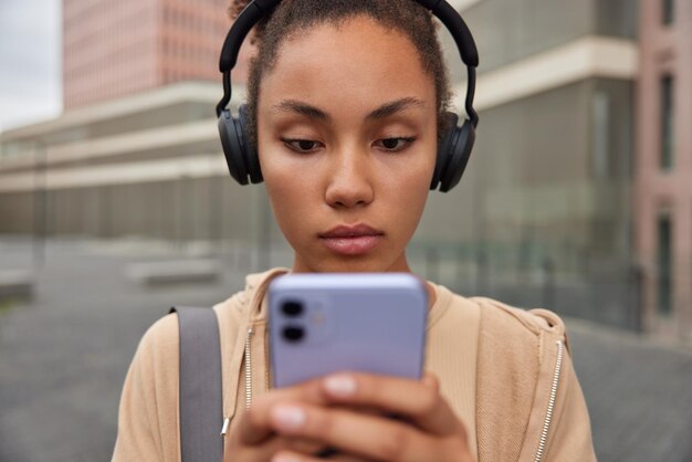 Horizontal shot of sporty woman listens music via headphones holds mobile phone concentrated into screen wears hoodie rests after fitness training rests after practicing yoga leads active lifestyle