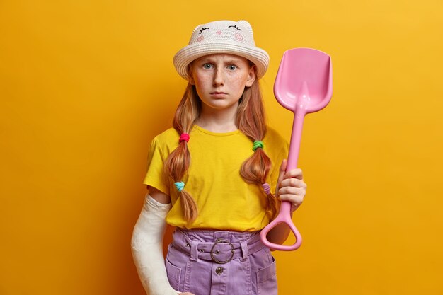 Horizontal shot of strict serious redhead girl plays in sand, holds pink plastic shovel, wears fashionable clothes, has broken arm bandaged in cast after doing dangerous sport, isolated on yellow wall