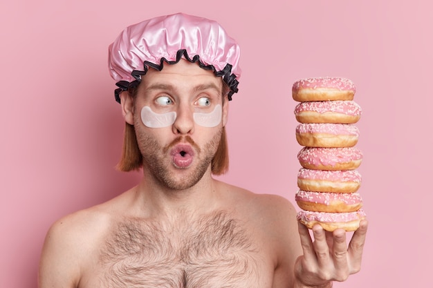 Free photo horizontal shot of stunned european man applies moisturizing pads under eyes keeps mouth opened stares at pile of delicious glazed doughnuts poses with bare shoulders against pink studio wall
