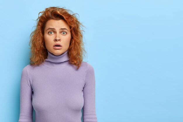 Horizontal shot of stunned woman looks with scary expression, has green eyes and wavy red hair