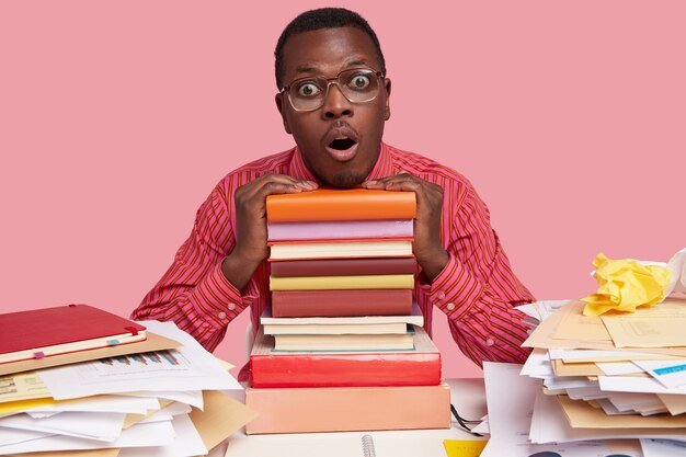 Horizontal shot of stupefied black man has scared expression, keeps both hands on textbooks