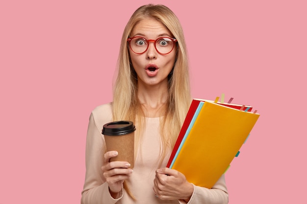 Horizontal shot of stupefied blonde woman in red framed eyewear, carries disposable cup of coffee, folders with stickers