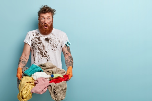 Horizontal shot of stupefied housekeeper carries laundry basket, has dirty clothing