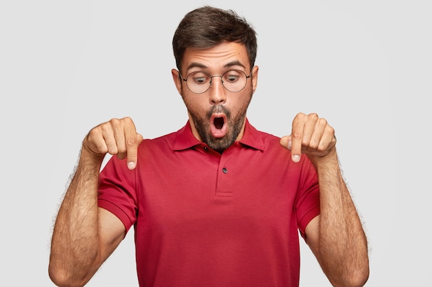 Free photo horizontal shot of stupefied young man looks surprisingly and indicates down, being stunned by something unbelievable, wears casual bright red t-shirt, stands against white wall