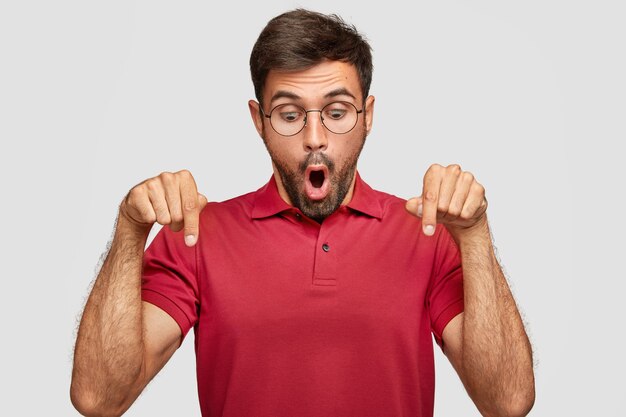 Horizontal shot of stupefied young man looks surprisingly and indicates down, being stunned by something unbelievable, wears casual bright red t-shirt, stands against white wall