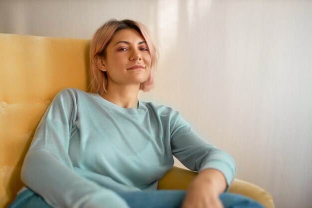 Horizontal shot of stylish pretty girl with bob pink hairstyle and nose ring sitting comfortably in armchair, looking at camera with carefree relaxed smile.