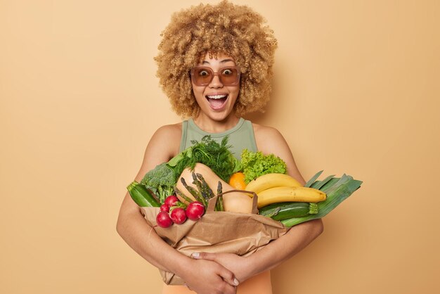 Horizontal shot of surprised amazed curly haired young woman embraces fresh vegetables and fruits wrapped in paper reacts to something unexpected carries grocery isolated over brown background