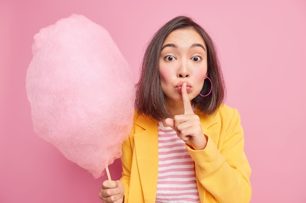 Horizontal shot of surprised Asian woman makes silence gesture asks to keep her secret presses index finger to lips holds tasty appetizing cotton candy wears stylish clothes. Hush dont spread rumors
