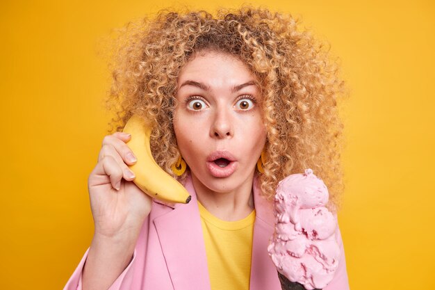 Horizontal shot of surprised curly woman stares bugged eyes holds breath keeps banana as if telephone near ear eats tasty ice cream during hot day gets too much calories has unhealthy nutrition