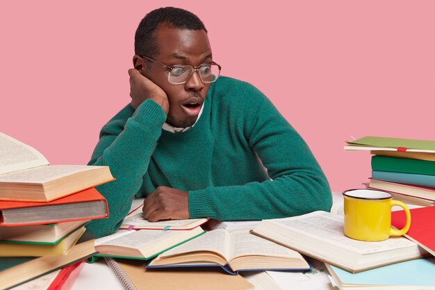 Horizontal shot of surprised dark skinned man wears optical glasses, focused with frightened expression at opened textbook