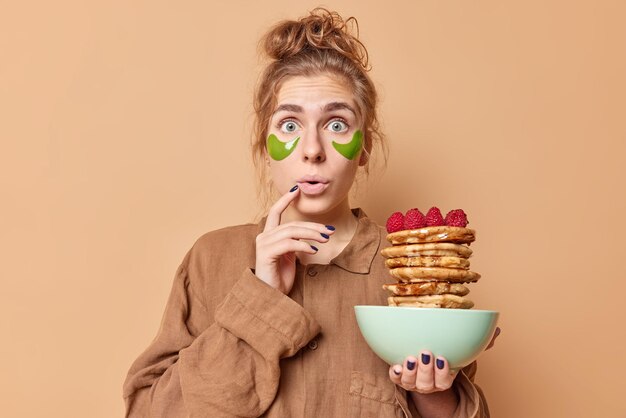 Horizontal shot of surprised emotional woman stares bugged eyes at camera applies green moisturising patches under eyes poses with bowl of pancakes dressed in pajama isolated over beige wall