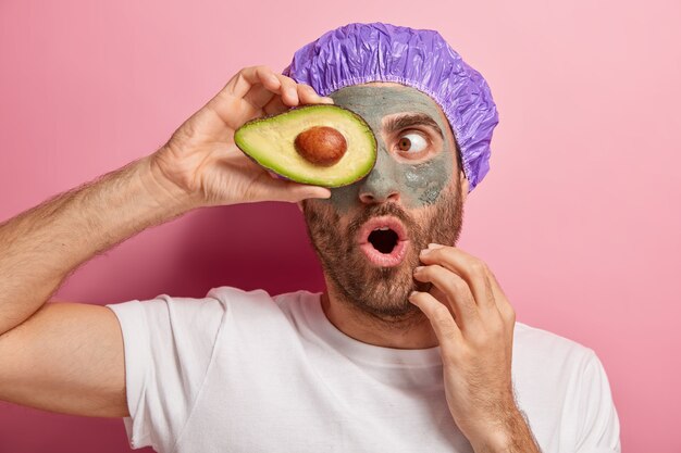 Horizontal shot of surprised European man keeps mouth opened from wonder, covers eye with slice of avocado, dressed in casual wear, shower cap