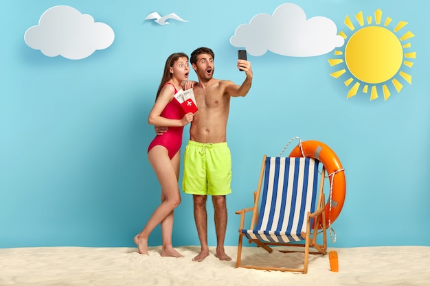 Colpo orizzontale di coppia femminile e maschile sorpreso piace trascorrere le vacanze estive al passaporto di spettacolo luogo resort con biglietti d'imbarco alla fotocamera del telefono cellulare fare selfie sulla spiaggia sopra priorità bassa blu